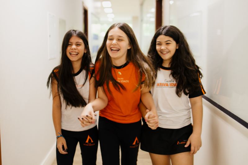 Três alunas do 6º ao 9º Ano de mãos dadas no corredor